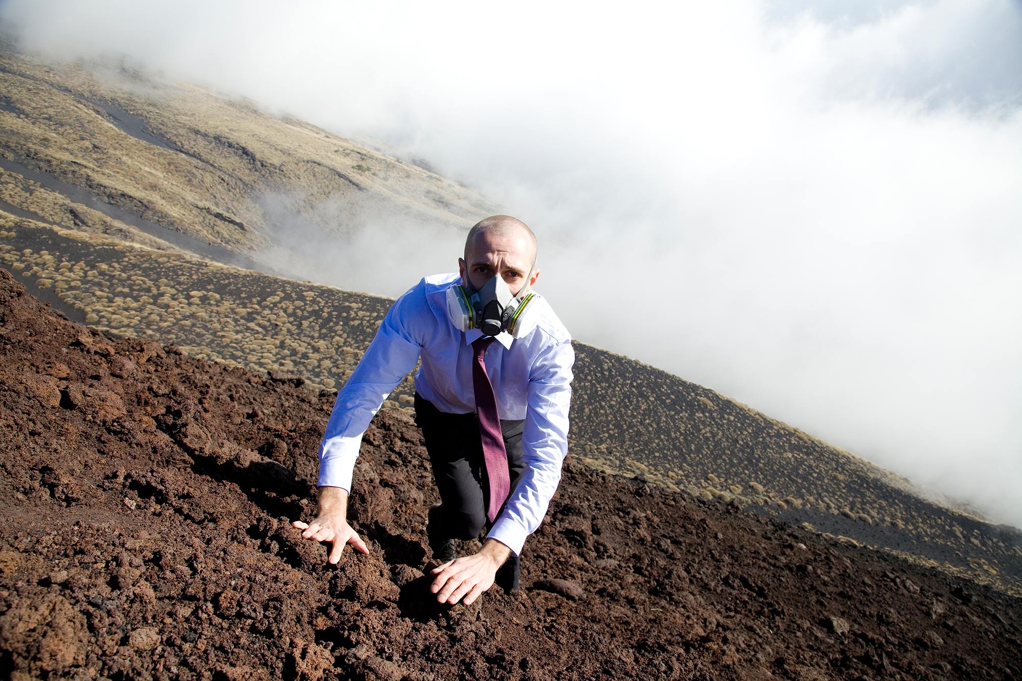 Matthias Mollner in a business dress and a gas mask climbs the hillside of a vulcano
