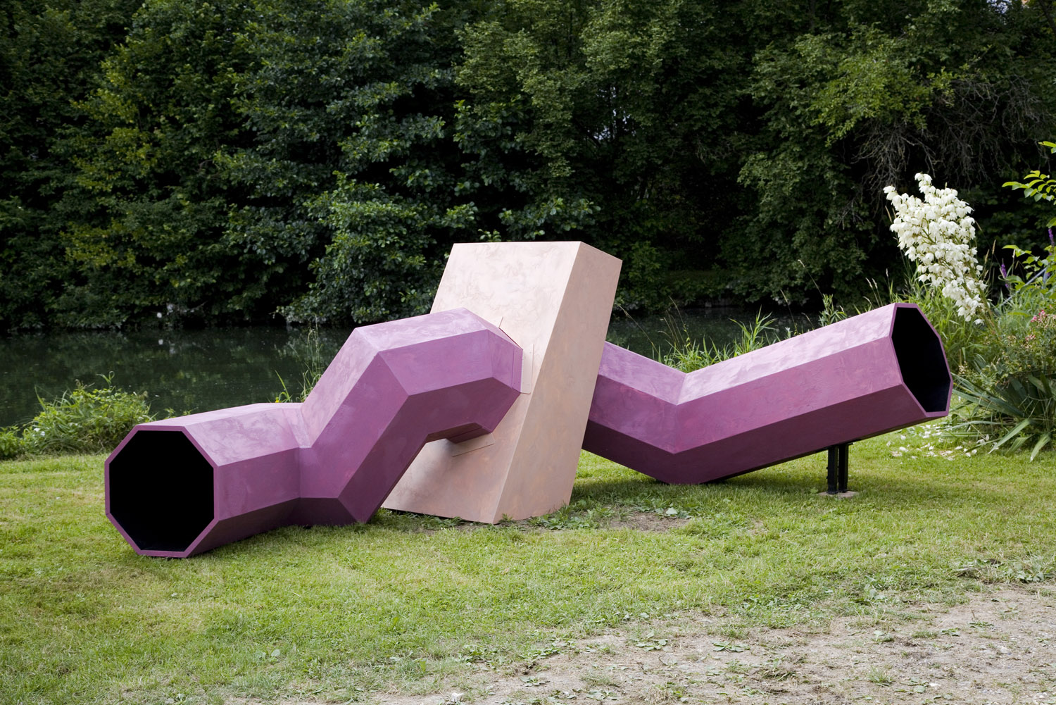 A winding pink and beige painted wormhole performance sculpture made of wood lies on a lawn next to a pond